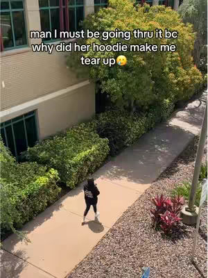 A high-angle shot captures a person in a black hoodie walking on a sidewalk, surrounded by greenery and a building. Text overlays the image. The person is walking away. The building has windows with a red-colored trim.