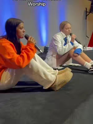 Two young women are seated on a dark surface, holding microphones and appearing to be singing or performing. The woman on the left wears an orange sweatshirt and the woman on the right wears a white jacket. The background is a blurred gradient of blue.