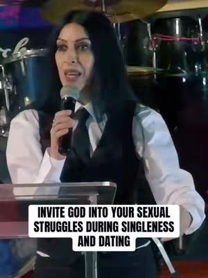 A woman with long black hair, wearing a white collared shirt, a black vest and tie, is holding a microphone and speaking. In the background, there are drums and other musical equipment. A white rectangular box with text overlay is at the bottom of the screen.