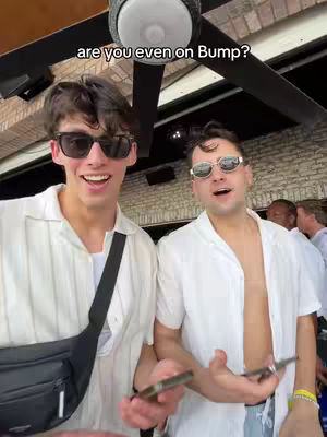 Two young men in casual attire are smiling at the camera while holding phones, with a background of an outdoor setting, including a ceiling fan.
