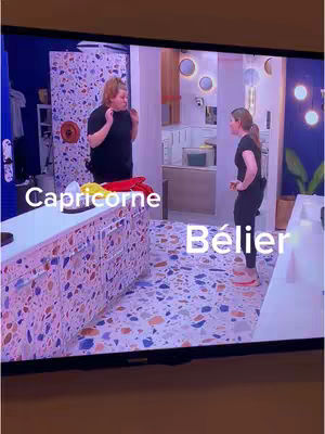 The cover image displays a scene captured from what appears to be a television screen. It shows two women in a domestic setting: one is standing with hands on hips, wearing a black top and pants, and the other is gesturing with hands, wearing a black top and skirt. They appear in a kitchen-like space with cabinets and flooring. Text overlays label the women as 'Capricorne' and 'Bélier'. The background includes kitchen elements and lighting. The composition suggests a scene from a video.