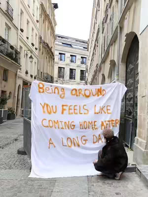 A person positioned in front of a large white sheet hanging in a narrow alleyway with European-style buildings on both sides. The sheet has text in what appears to be orange paint, reading Being around You Feels like Coming home after a long day. The person is squatting and looking at the sheet.