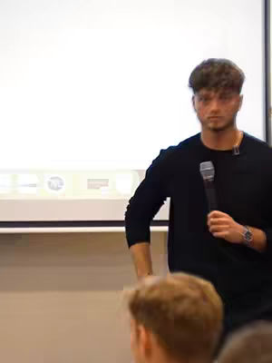 A man in a black shirt holds a microphone, standing in front of a white screen or projector. Other individuals are visible in the foreground, suggesting a conference or presentation setting.