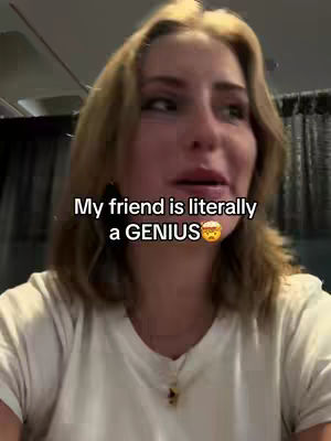 A young woman with blonde hair and fair skin appears in a close-up shot, slightly off-center. The soft lighting highlights her face and the white t-shirt she is wearing. The background is blurred, but it seems to be indoors, with dark curtains or a similar element visible.