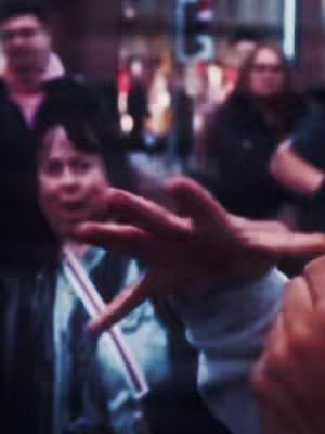 The cover image is a blurred street scene, likely taken during the day. The primary focus is on a woman in the midground looking shocked, with an open mouth, a hand in front of her and a hand's fingers fully stretched to the foreground, with a soft focus. She is wearing a white top with suspender straps. The background fades into a blurry crowd, with other people and building outlines discernible. Colors suggests daylight with a slight blueish tone, and the overall feel is one of a candid shot.