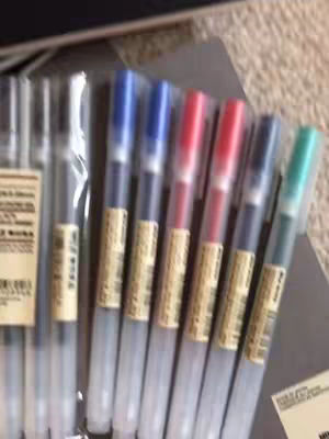 The cover image shows a close-up of a collection of Muji pens. The pens are arranged in a row, with their transparent barrels and colored caps and ink on display. They are partially inside of a clear plastic container.