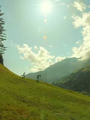 A scenic image of two silhouetted figures on a green grassy hill, beneath a bright sun in a lightly clouded sky. Mountains are visible in the distance.