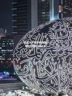 A night shot of a futuristic, abstract building with glowing white text. The building's design is intricate, and the overall composition is striking against a dark background of city lights. The architecture appears modern, with a reflective silver surface catching the light.