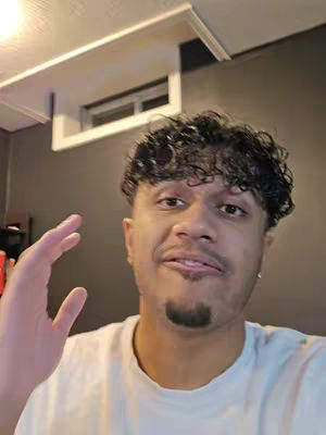 A man with dark curly hair and a goatee is in a close-up shot, gesturing with his hand. The background features a dark wall and a section of the ceiling.