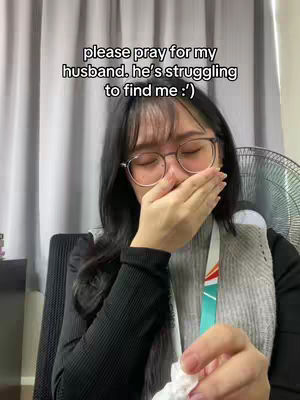 A woman sits in front of a window curtain, covering her mouth with her hand. She wears glasses and a black long-sleeved shirt. Text overlaid at the top says, "please pray for my husband. he’s struggling to find me :)"