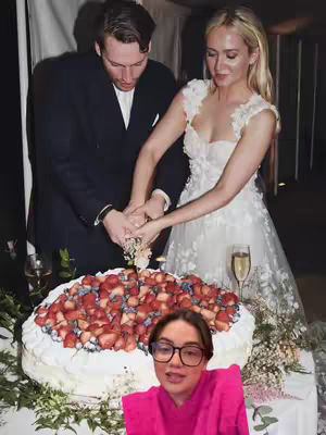 A wedding scene with a bride and groom cutting a cake. The bride is wearing a white wedding dress, and the groom a black suit. In the foreground a woman with glasses reacts in front of the cake, which is decorated with strawberries.