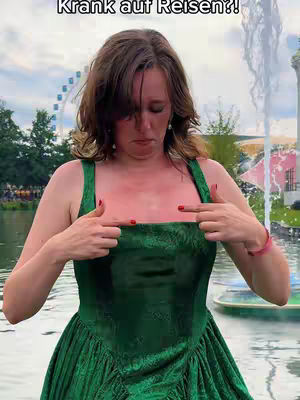 A woman in a green dress stands facing the camera outdoors. She is pointing at the reddened area on her chest. In the background, a fountain and a Ferris wheel are visible, suggesting a park or recreational area.