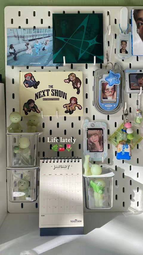 A vertical image showcasing a white pegboard filled with various items. There are multiple small containers and shelves holding figurines and small toys. Photos are placed in decorative frames or holders. A calendar is visible with the month of January displayed. Various stickers and decorative elements also adorn the board. The text 'Life Lately' is placed near the calendar, and the words 'THE NEXT SHOW' is on the calendar.