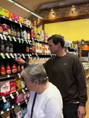 A medium shot captures a man and a woman in a grocery store aisle, looking at products on the shelves. The man is slightly taller and appears to be pointing at something with his right hand. The scene is brightly lit, with a display of various grocery items in the background.