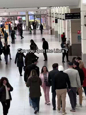 An image taken in a public space, likely a mall or airport, with numerous people walking in various directions. The composition is from a higher angle, looking down a hallway. The color palette is fairly muted, with a focus on natural and neutral tones. The foreground is occupied by a crowd of people, while the background shows more people and architectural details. The lighting seems to be indoor, with a lot of artificial light.