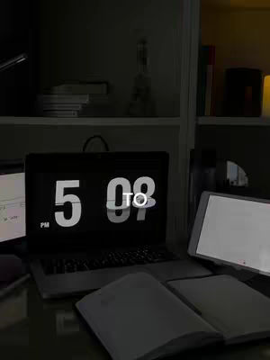 A dimly lit scene with a laptop prominently displaying the time '5:08 PM,' positioned on a sleek desk. A notebook and a tablet are next to the laptop. Shelves containing books are partially visible in the background, adding to the study environment.
