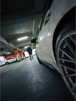 A low-angle shot inside a parking garage, dominated by a white car on the right. A man is walking toward the camera in the center. The background features orange and white pillars and other parked cars.