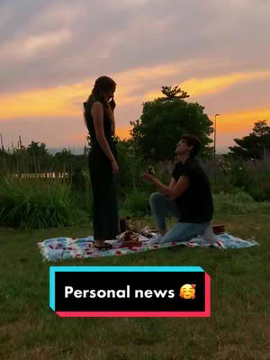 Two women are in an outdoor setting, one kneeling and presenting a ring to the other who stands facing her. They're on a patterned blanket on green grass, with the backdrop of a cloudy sky. A text overlay at the bottom says 'Personal news' with an emoji.