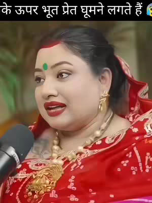 A woman with fair skin and dark hair is centered in the frame, partially obscured by a microphone. She wears a bright red saree with intricate patterns and a gold necklace. The background is out of focus, suggesting an indoor setting. Additional text is overlaid at the top.