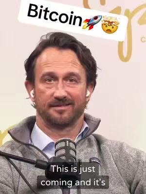 A man with dark hair wearing a gray zip-up hoodie sits in front of a microphone. Text bubbles with the word 'Bitcoin' and associated emojis are above his head. The background is a blurred light color.