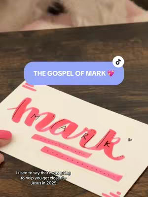 A handwritten card that reads 'THE GOSPEL OF MARK' in a light purple rounded rectangle. The word 'MARK' is written in red and pink. The image is shot from an above angle, with the corner of the card in focus. The background is brown.