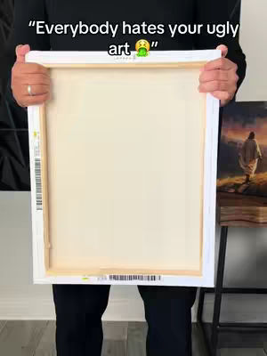 A person holds a stretched canvas, viewed from the back, against what appears to be a white wall. The canvas is blank, with its wooden frame visible. The person's lower body and arms are also visible. To the right, a painting is visible, with a religious theme. The image is well-lit and neutral in tone.