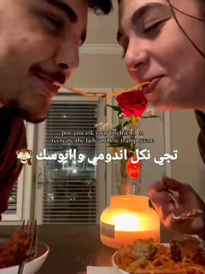 A couple is shown from the chest up, each eating a strand of spaghetti, drawing them closer. The setting appears to be a romantic dinner setting with a candle and a flower in a vase. The background is blurred, suggesting a cozy interior.