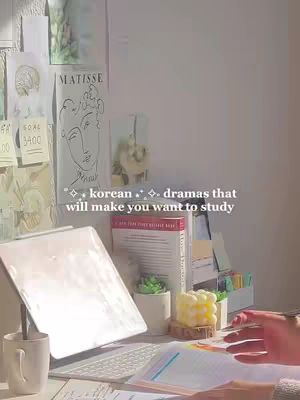 The image shows a cozy and well-lit study space. A laptop is open on a desk, with a mug, books, stationary and candle are also on the desk. On the background, there are some posters on the white wall.