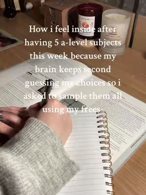 A desk is shown with an open notebook and a textbook. A hand is writing in the notebook. On the desk are a candle and various stationery supplies. The overall composition suggests a study environment, likely a student's workspace.