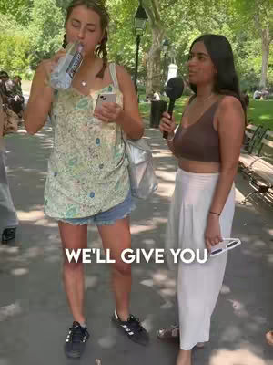 Two young women are standing outside in a park and near a sidewalk. The woman on the left is drinking from a water bottle and holding a phone, while the woman on the right is holding a microphone. A text overlay in the bottom of the cover image says: “WE’LL GIVE YOU”.
