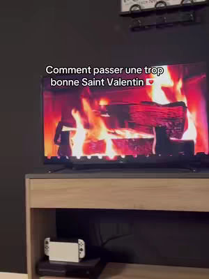 A medium shot shows a flat-screen TV displaying a fireplace scene, with flames and burning logs. Text overlays the screen. Below the TV is a wooden cabinet containing a game console and other items. The background is a simple black wall.