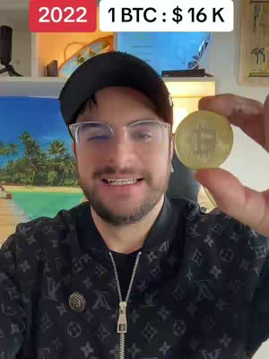 A man wearing glasses and a black cap holds a Bitcoin coin towards the camera, with a background of a shelf and a view of the ocean from a window. A text overlay at the top shows '2022 1 BTC : $16 K'.