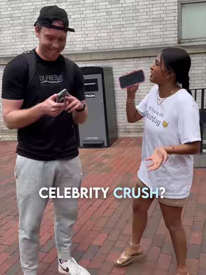 A man and a woman standing outside, likely on a college campus, both holding phones and smiling. Text on the image asks about a celebrity crush. The man wears a black t-shirt and a hat, while the woman wears a white t-shirt with text. The background includes a brick wall, a trash can, and some greenery.