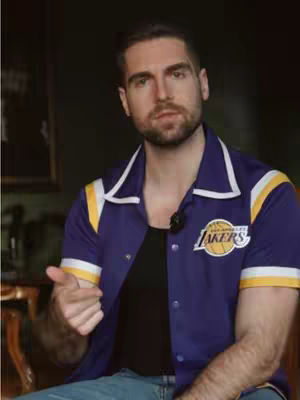 A man in a Lakers jacket, pointing his finger towards the viewer. The background is dark and out of focus.