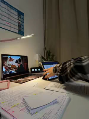 A well-lit desk scene with a laptop displaying a cartoon, a tablet, and scattered papers suggesting a study session. The composition includes a calendar, a clock, and a hand interacting with the tablet, with cozy lighting.