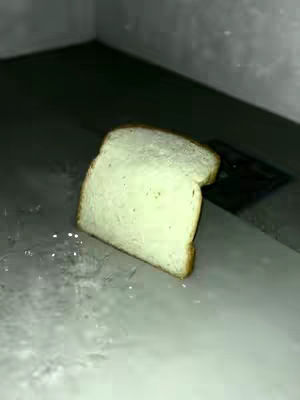 A slice of bread is shown resting at a diagonal on what appears to be a metal surface. The lighting is dim, creating shadows and a sense of depth. The background is out of focus, suggesting a close-up shot.