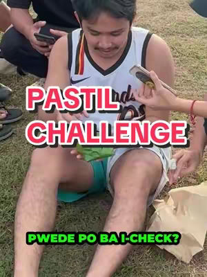 A man in a white and black jersey sits on the grass, looking down at a piece of food in his hands. Overlay text in pink and white font reads 'PASTIL CHALLENGE' and 'PWEDE PO BA I-CHECK?'