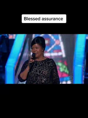 A woman with short, styled black hair is holding a microphone and singing. She is wearing a black and patterned top with a dark background, possibly on a stage. Above her in a rectangle is the text overlay 'Blessed assurance'.