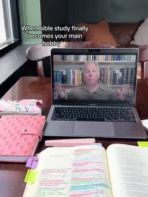 A close-up shot of a laptop displays a video call with a man giving a sermon or lecture on a background of books, papers, and other study materials are spread across the desk.