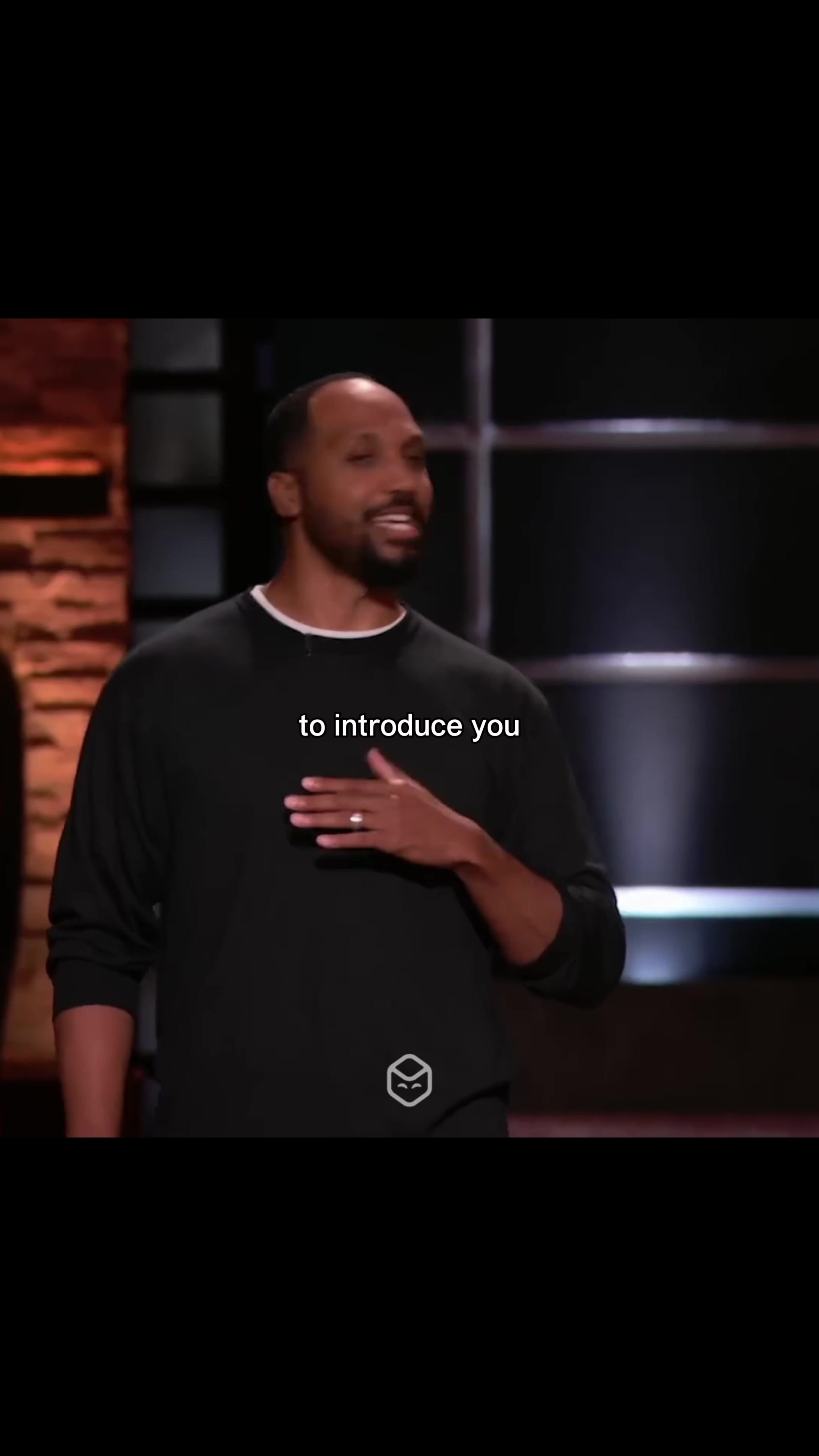 The image depicts a man in a black long-sleeved shirt standing on what seems to be a stage. Text overlayed at the bottom of the image reads 'to introduce you'. The background is dark and out of focus, and a logo is visible on the man's shirt.