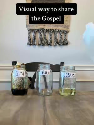 The cover image shows three glass jars on a wooden table, each labeled with a word: "SIN," "YOU," and "JESUS." The jar labeled "SIN" contains a dark liquid, while the "JESUS" jar holds a yellowish liquid. The table is set in front of a white wall with a decorative woven hanging above.