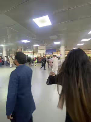 An airport interior is shown. Two people are in the foreground; one is a person facing away with long dark hair, and is extending their arms out toward the right side of the frame. The other person a is male figure, with a blue suit, walking towards the camera. People are in the background as well, some are pushing trolleys and the floor is a light beige color.