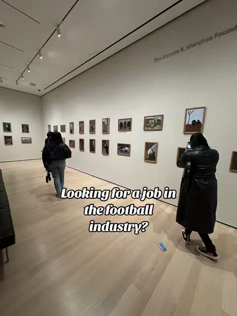 The image shows a brightly lit art gallery with a light-colored floor and white walls. Multiple framed artworks are arranged on the walls. A person wearing a black coat stand in the frame with their back towards the camera, using phone. Another two people walk away in the background. Overlaid text is present.