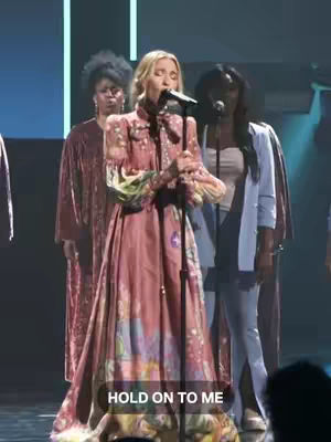 A woman is singing into a microphone on a stage, wearing a long, patterned dress. Two women stand behind her as backup singers. The lighting is dim, with a spotlight on the singer. The background is dark.
