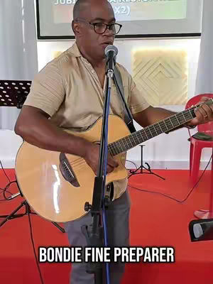 A man with glasses is playing an acoustic guitar and singing into a microphone. He's in a brightly lit room with a red floor and a white background.