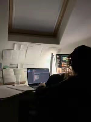 Low-lit image of a person sitting in front of a laptop and a desktop monitor, likely in a home office or study area. The person's face is obscured. The laptop screen is visible, displaying what appears to be code. Papers are pinned to the wall. A desk lamp provides a warm glow.