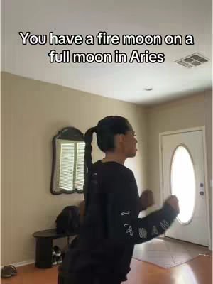 A woman with dark hair in a ponytail is dancing in a hallway. She is wearing a black sweater, the background includes a mirror, a table, and a door.