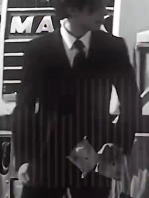 A black and white shot shows a man in a pinstripe suit and tie. He is slightly out of focus, and there is a partial view of a sign behind him that reads 'MA'. The composition is simple, with a focus on the man's upper body and the pinstripe of the suit providing vertical lines.