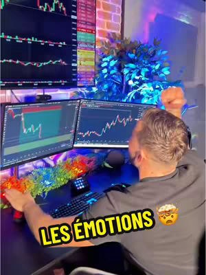 A person is sitting at a desk with multiple computer screens. The screens display financial data charts with lines and numbers. The person is facing away from the camera with arms raised up, as if in reaction to something on the screen. A text overlay with the words "LES ÉMOTIONS" and a small emoji is present.
