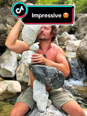 A man with curly hair and no shirt is balancing rocks, surrounded by a natural setting with rocks and water, has text overlayed on the top right.
